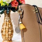 silver metal badminton racket keychain with a small pink and white plastic shuttlecock attached, Displayed on Tan handbag as bag charm against a plain beige background.


