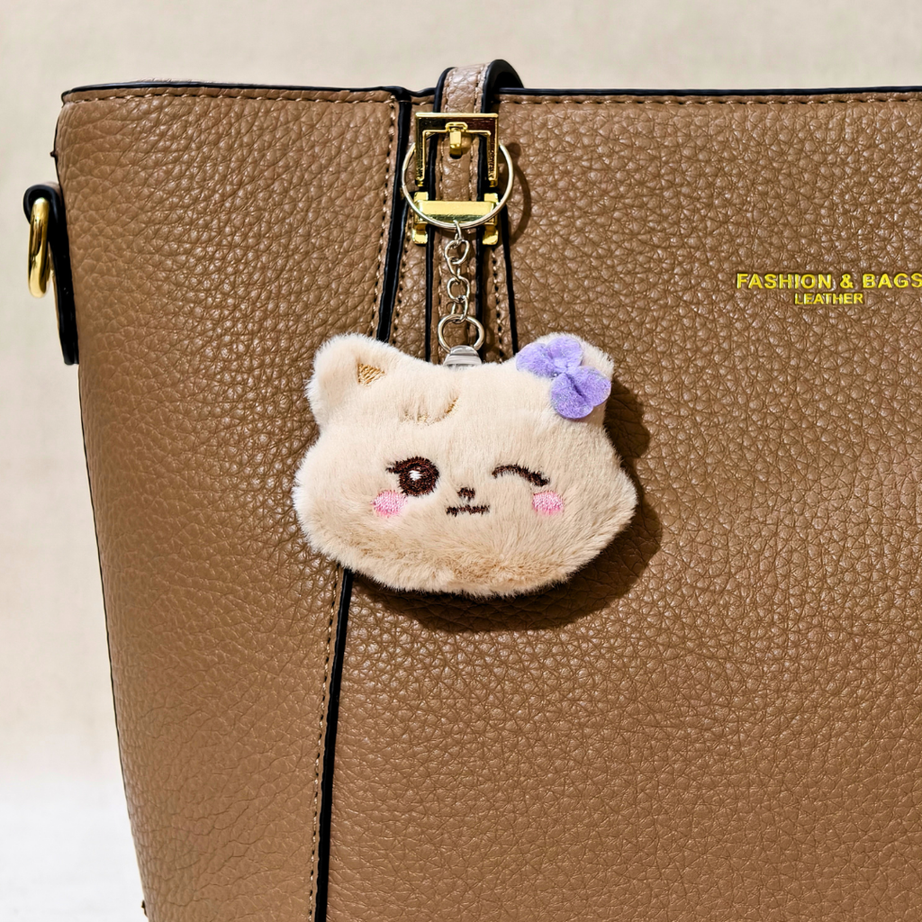 A close-up of a brown textured leather handbag with a cute plush cat keychain charm hanging from the gold-tone buckle. The cat is cream-colored with a purple flower on its head, winking with pink cheeks.