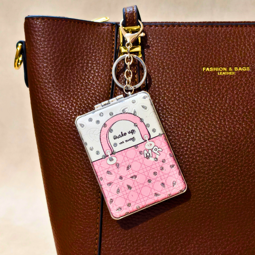 A pink and white handbag-shaped keychain charm with a quilted pattern and the words 'Make up is not sing' hanging from a gold key ring, shown against a brown textured leather bag.