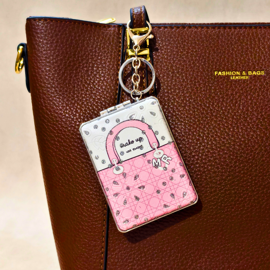 A pink and white handbag-shaped keychain charm with a quilted pattern and the words 'Make up is not sing' hanging from a gold key ring, shown against a brown textured leather bag.