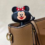 A black silicone AirPods case featuring a Minnie Mouse face design with her signature red and white polka-dot bow, shown resting on top of a brown textured leather handbag.
