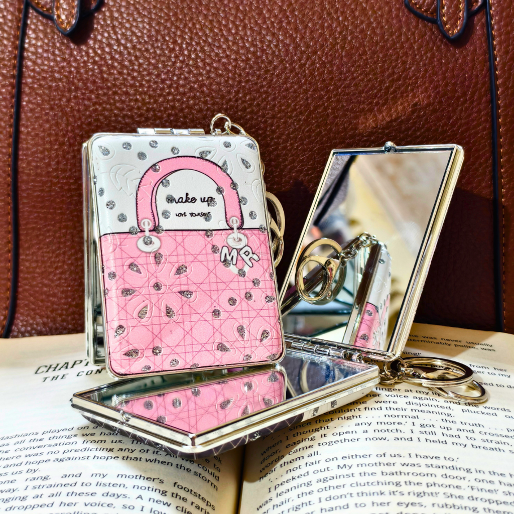 A pink and silver quilted handbag-style compact mirror with a built-in ring, displayed open on an open book with a brown leather bag in the background.