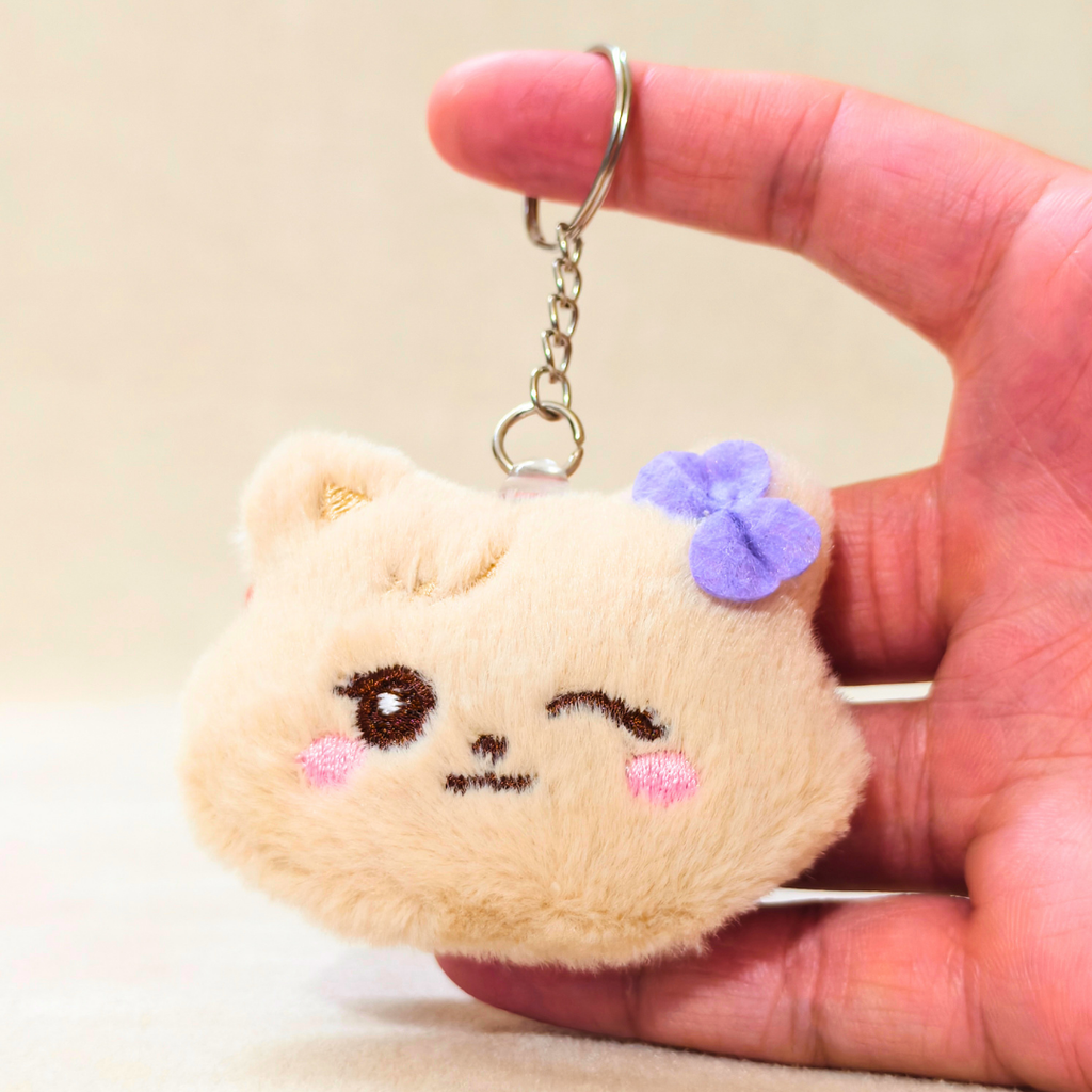 A hand holding a small plush keychain shaped like a winking beige cat face with pink cheeks, a purple flower accent, and a silver key ring.