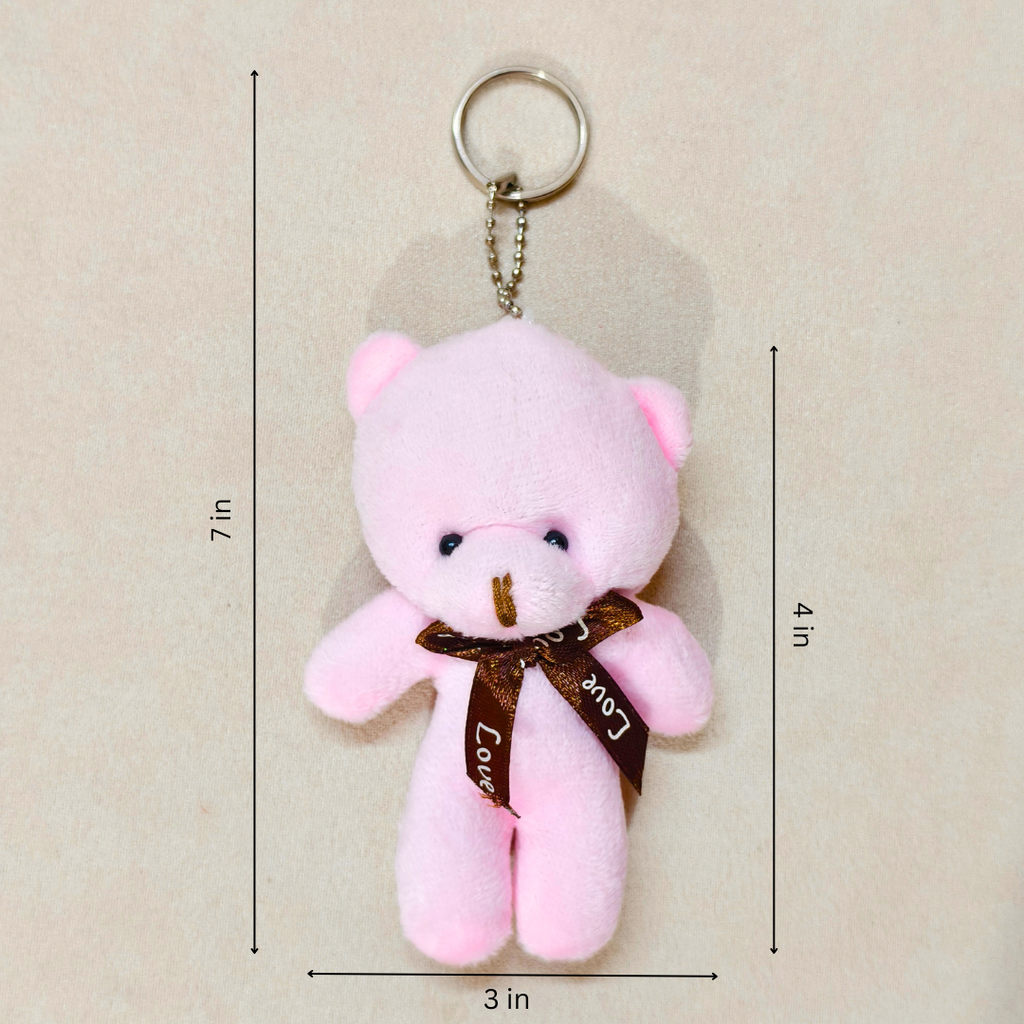A pink plush teddy bear keychain with a brown ribbon and metal key ring, shown on a light beige background with dimension lines indicating 7 in height, 4 in width, and 3 in depth.