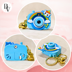Three circular product views of a small blue camera-shaped keychain with a rainbow, clouds, stars, cat-face detail, and gold bell keyring on a soft pink background. A stylized 'BV' logo appears in the top left.
