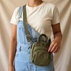 Person wearing blue overalls and a white shirt holding a green bag against a beige background