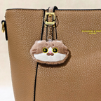 A plush keychain shaped like a small brown and white cat face, held in a person's hand. The keychain has yellow embroidered eyes with black pupils, a pink nose, and a silver metal key ring and chain Displayed on a brown handbag as bag charm on a beige background.