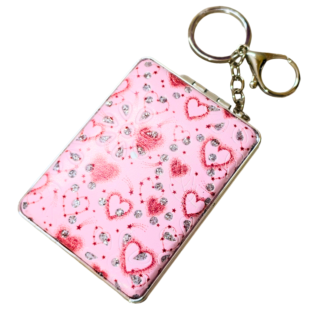 A pink rectangular card holder or compact-style case with a silver metal frame and attached gold-tone key ring and chain, decorated with red hearts and rhinestone-like sparkles on a white background.