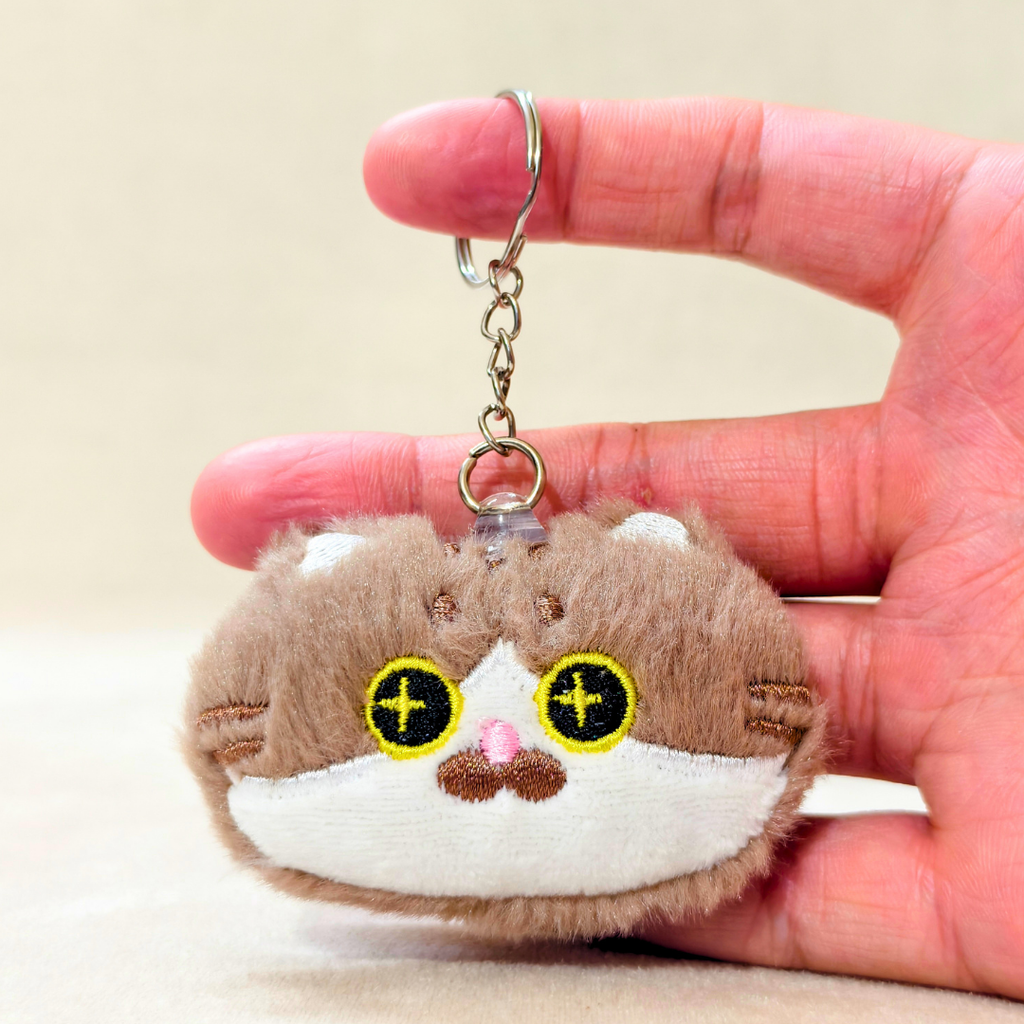A plush keychain shaped like a small brown and white cat face, held in a person's hand. The keychain has yellow embroidered eyes with black pupils, a pink nose, and a silver metal key ring and chain.