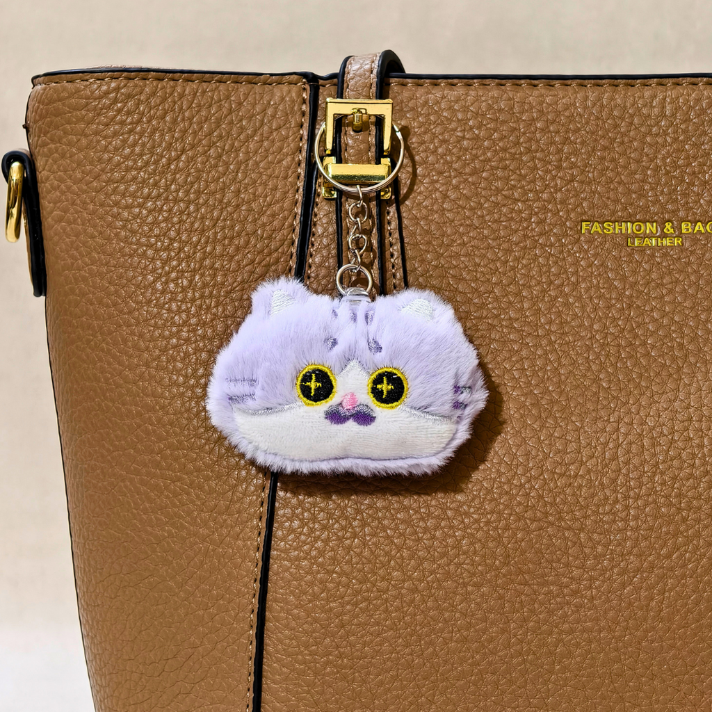 A plush cat-shaped keychain charm in light purple and white with embroidered yellow eyes and a small pink nose, shown on a  brown Hand Bag  With beige background .