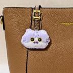 A plush cat-shaped keychain charm in light purple and white with embroidered yellow eyes and a small pink nose, shown on a  brown Hand Bag  With beige background .