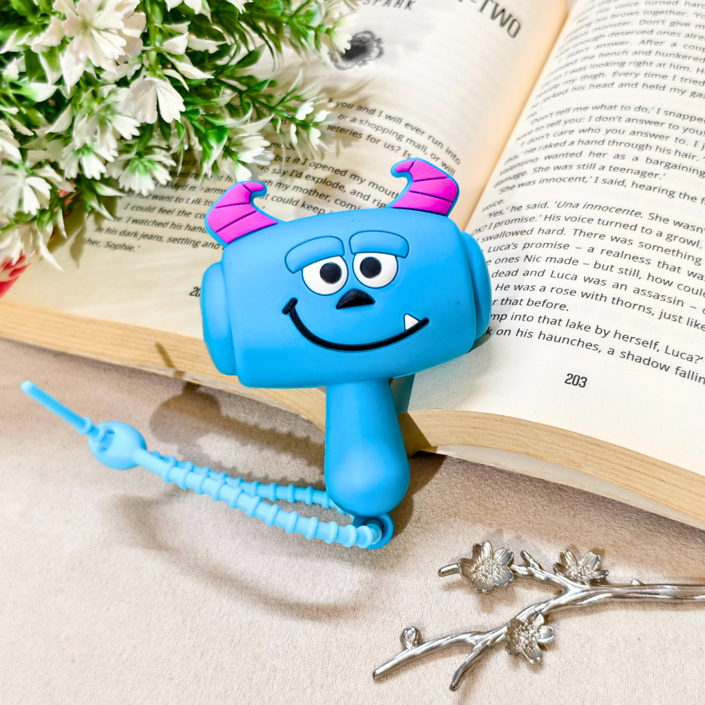 A blue monster-shaped clip-on bookend with pink horns, a smiling face, and a coiled blue string, styled on an open book with flowers and silver jewelry in the background.