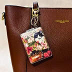 A brown textured leather handbag with gold hardware and the text 'FASHION & BAGS, LEATHER' on the side, featuring a hanging rectangular charm decorated with a floral print and two cats.