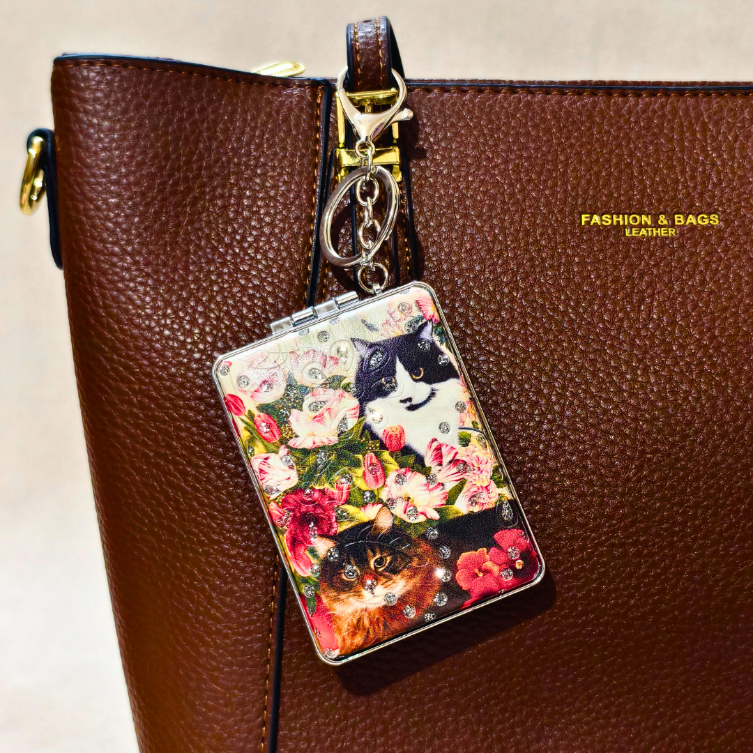 A brown textured leather handbag with gold hardware and the text 'FASHION & BAGS, LEATHER' on the side, featuring a hanging rectangular charm decorated with a floral print and two cats.