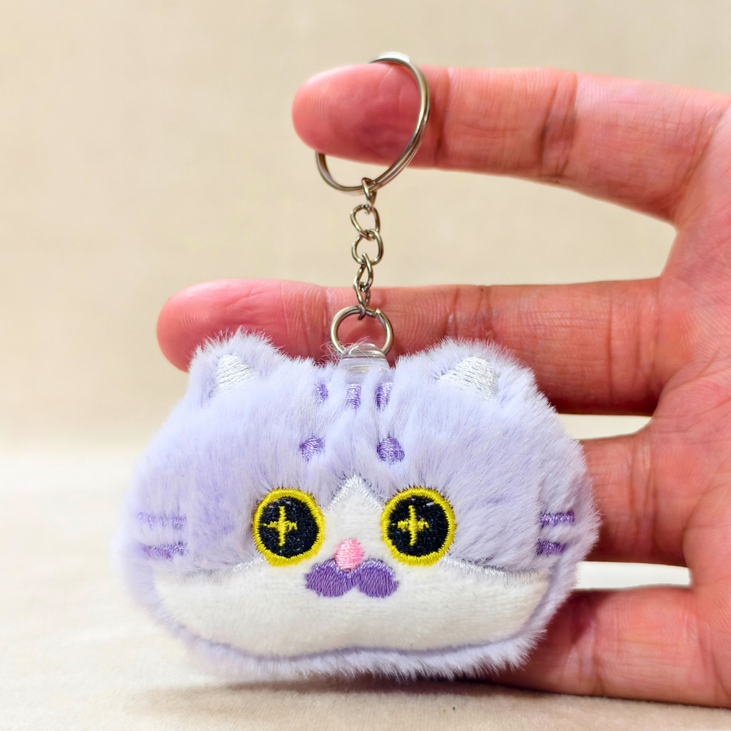 A small plush keychain shaped like a cat face in pale lavender and white, held in a person's hand. The keychain has yellow embroidered eyes with black crosses, a pink nose, and a silver metal key ring and chain.