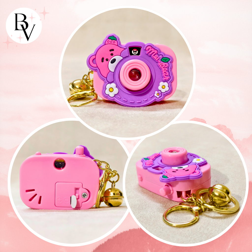 Three circular product views of a small camera-shaped keychain in pink and purple, with gold keys and a bell, shown on a soft pink background. A stylized 'BV' logo appears in the top left.