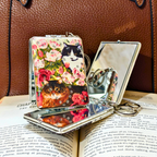 A silver metal compact mirror with a decorative floral and cat print case, displayed open on an open book with a brown textured surface in the background.