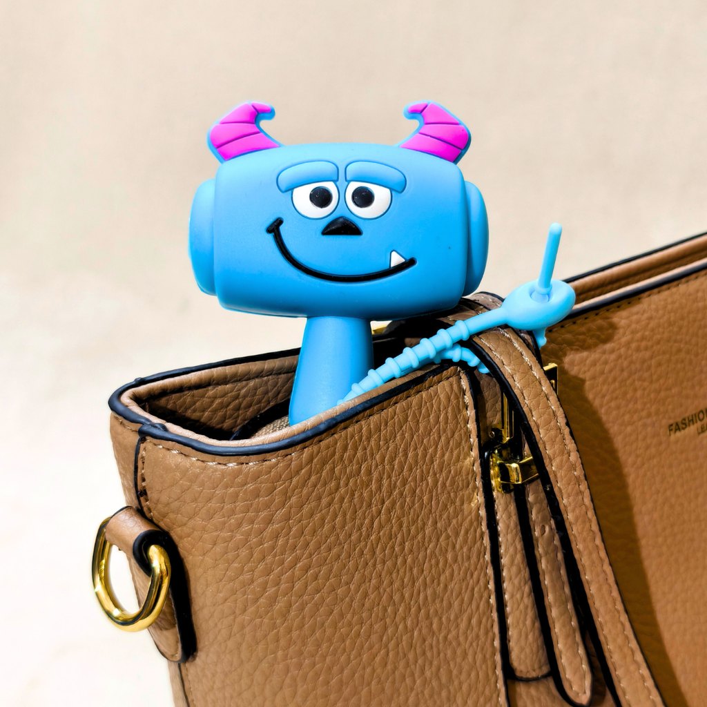 A blue cartoon monster-shaped zipper pull attached to a brown textured leather handbag, with the monster's horn and tail visible against a light beige background.