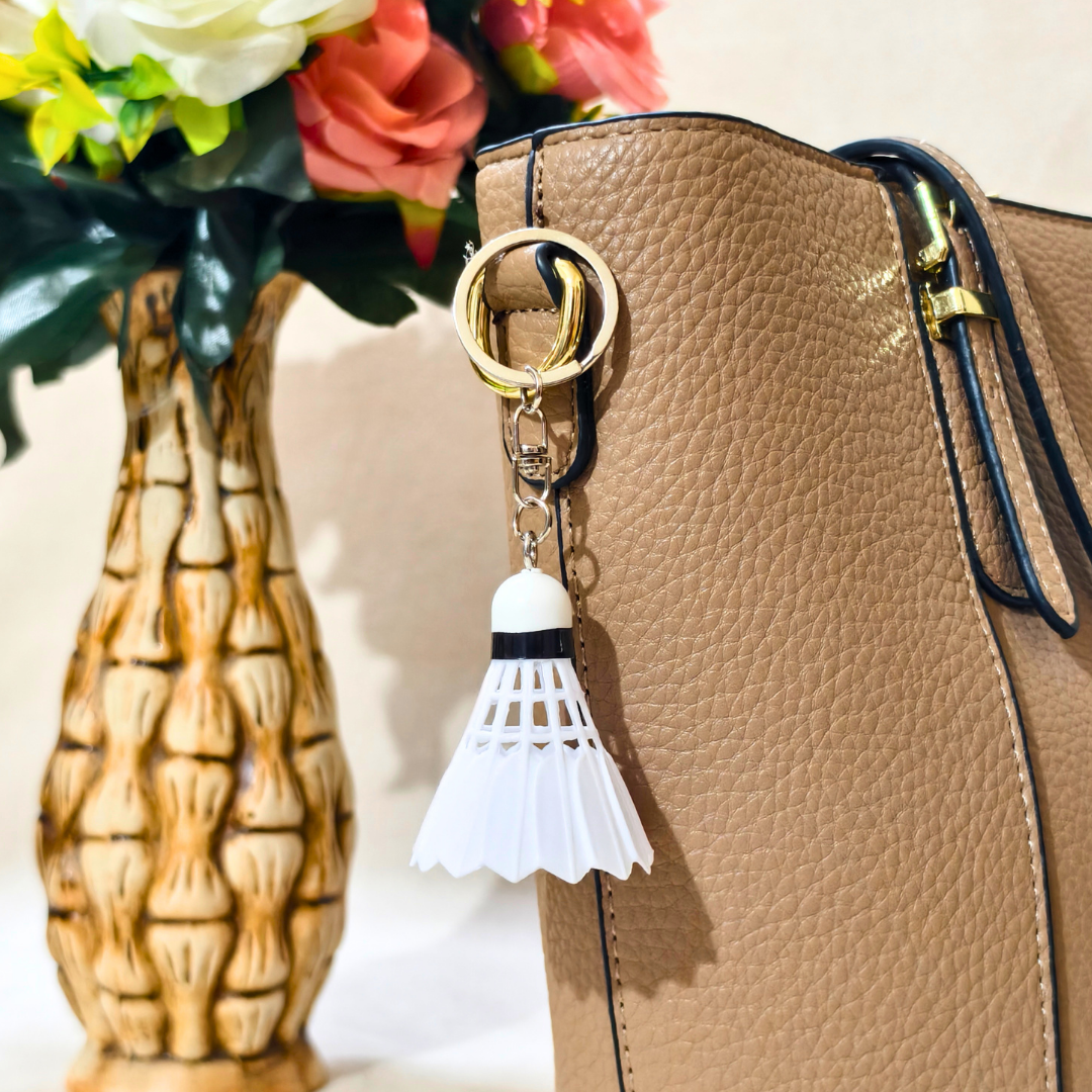 A white plastic shuttlecock keychain with a silver metal split ring and chain,  displayed on Handbag as Bag Charm. Shown on a beige textured background.