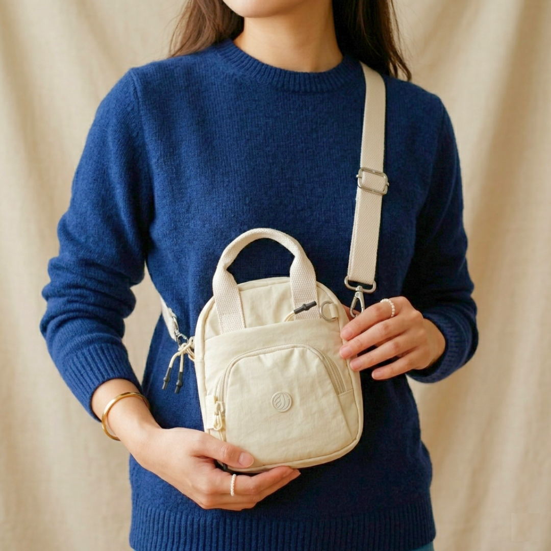 Person wearing a blue sweater holding a beige crossbody bag against a beige background