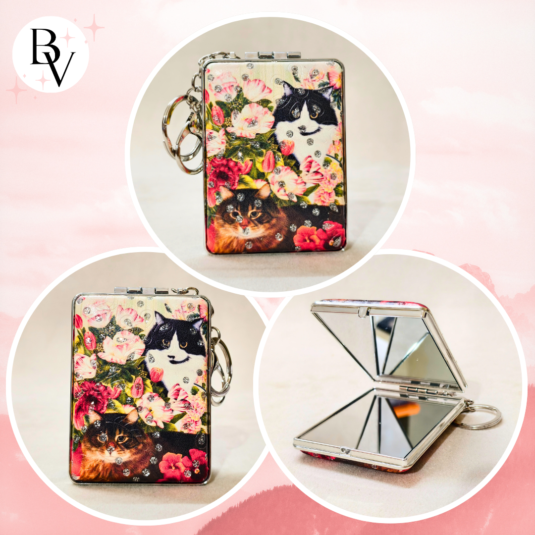 A compact folding mirror case with a floral print featuring cats and roses, shown closed and open on a soft pink background. The product has a silver-tone metal frame, keychain ring, and mirror inside.