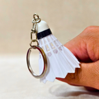 A white plastic shuttlecock keychain with a silver metal split ring and chain, held on hand shown on a beige textured background 