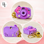 Three circular product views of a small purple toy camera keychain with a pink bear face, white flowers, and a gold bell keyring on a pale pink background. A stylized 'BV' logo appears in the top-left corner.