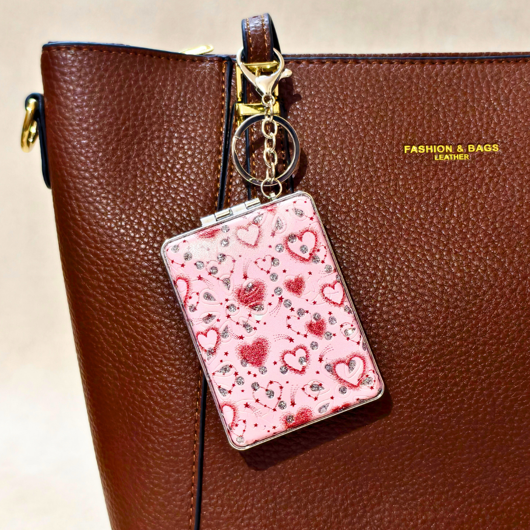 A pink heart-patterned card holder keychain with silver and red glitter details, attached to a gold key ring and strap, shown hanging from a brown textured leather bag.