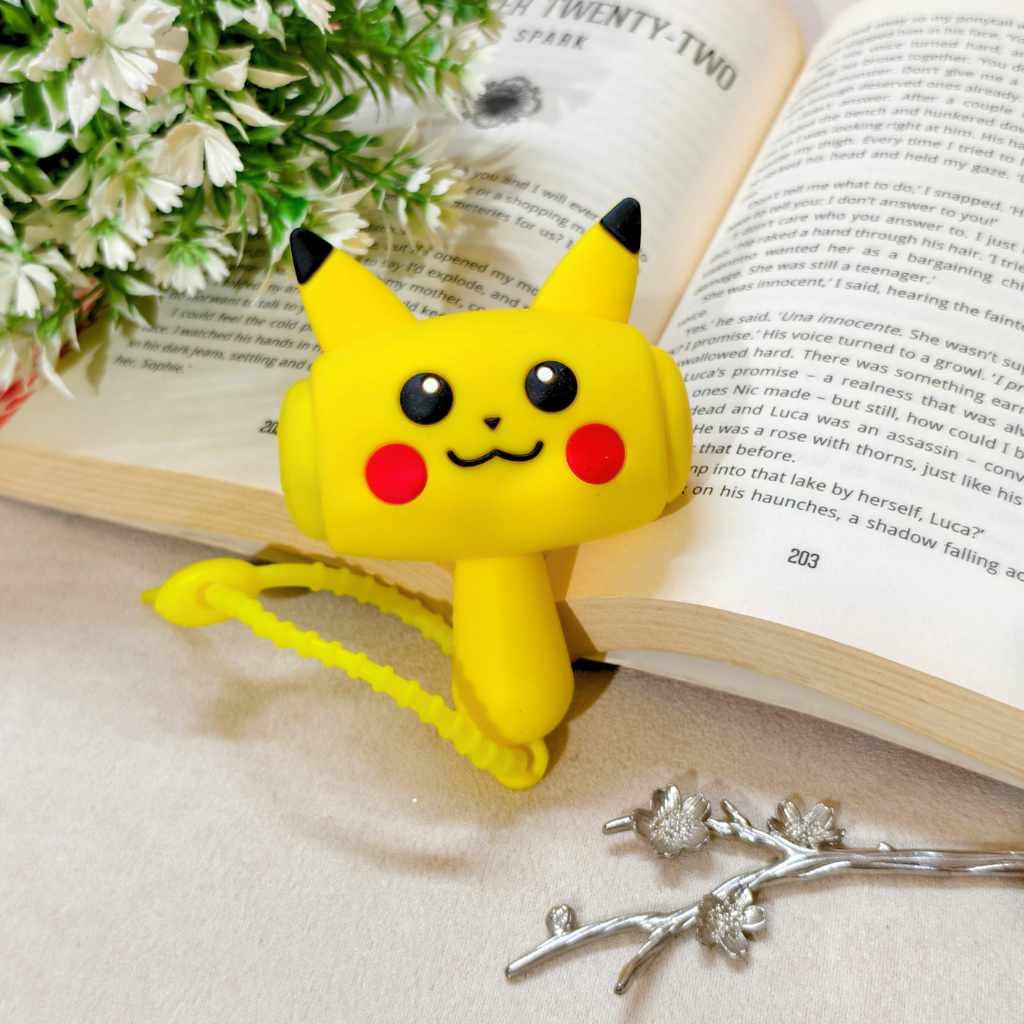 A yellow Pikachu-shaped book clip holding an open book on a light beige surface, styled with white flowers and silver branch accessories.