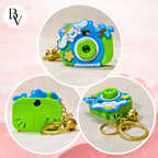 Three circular product views of a small camera-shaped keychain charm on a soft pink background. The charm is bright green and blue with a cartoon character face, gold keychain rings, and the word 'cinnamoroll' printed on the front.