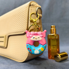 Keychain shaped like a cartoon donut with pink hair next to a gold handbag and perfume bottle on a gray background