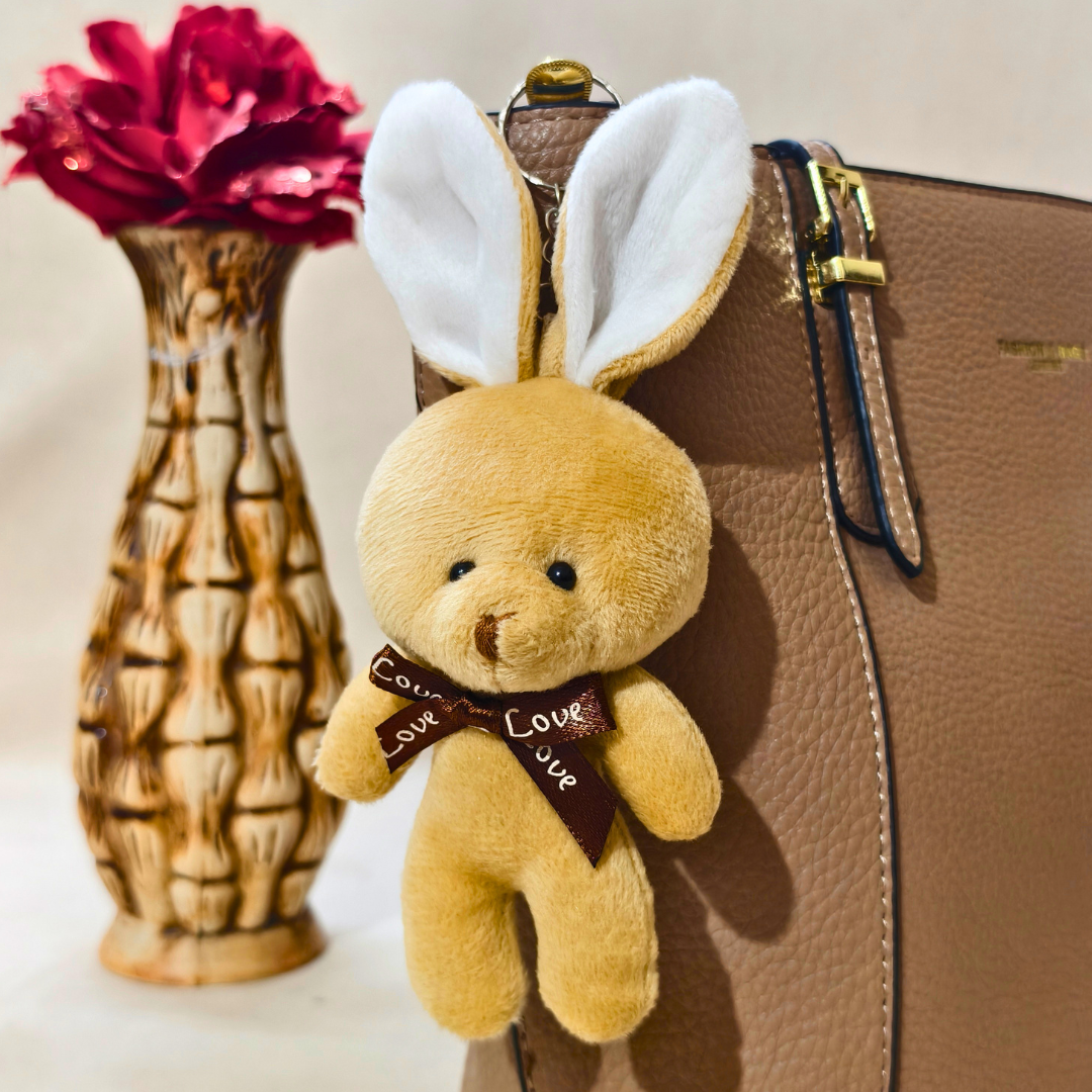 Small teddy bear with bunny ears attached to a brown suitcase next to a wooden vase with red flowers.