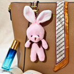 A pink plush bunny keychain with long ears and a small dark brown bow, hanging from a tan textured handbag. The handbag is styled with a patterned scarf and a blue perfume bottle nearby.