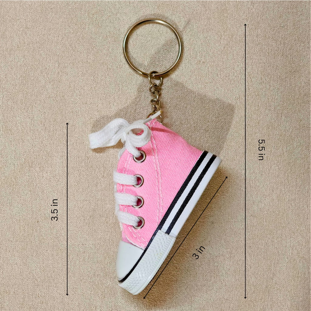 Pink canvas high-top shoe keychain with white laces, white rubber toe cap and sole, and a gold metal key ring, shown on a beige textured background with dimension lines and measurements.