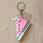 Pink canvas high-top shoe keychain with white laces, white rubber toe cap and sole, and a gold metal key ring, shown on a beige textured background with dimension lines and measurements.