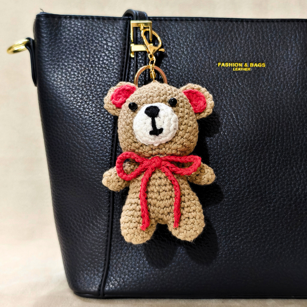 Crocheted teddy bear keychain with a red bow on a black leather bag.