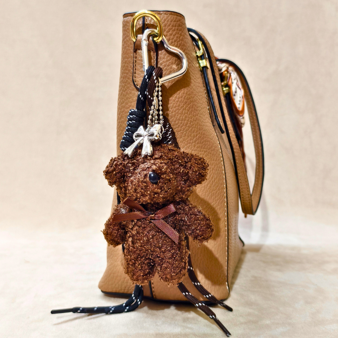 Brown teddy bear keychain attached to a beige handbag on a light beige background