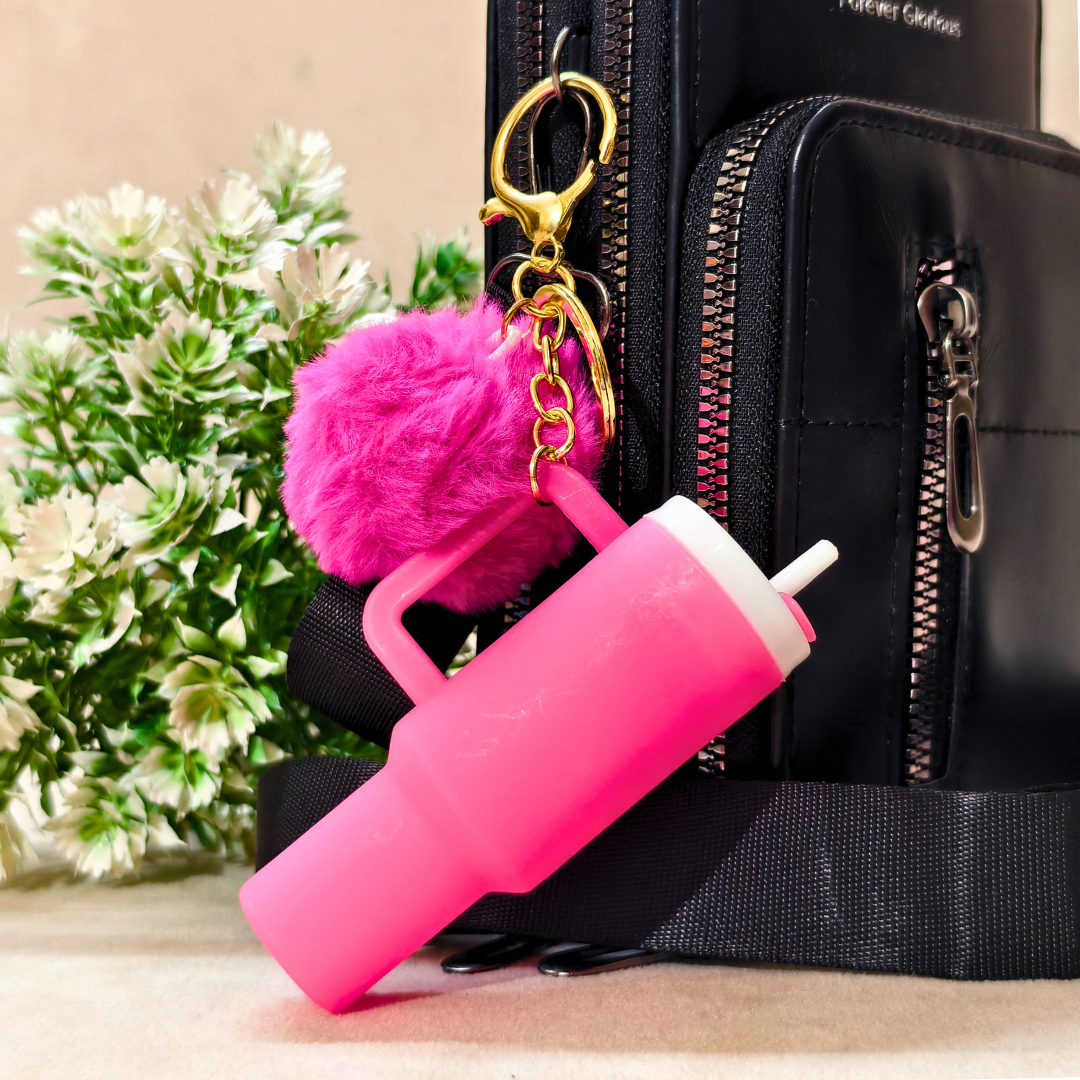 A bright pink reusable bottle with a handle and straw, attached to a gold key ring and a fluffy pink pom-pom, shown in front of a black zippered bag with decorative flowers in the background.
