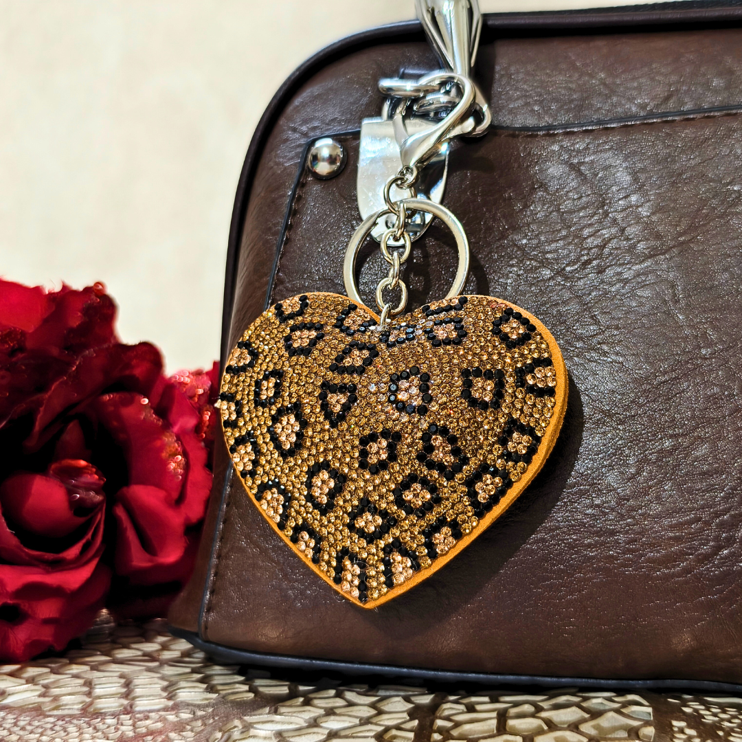 Heart-shaped keychain with leopard print pattern attached to a brown leather bag next to a red rose.
