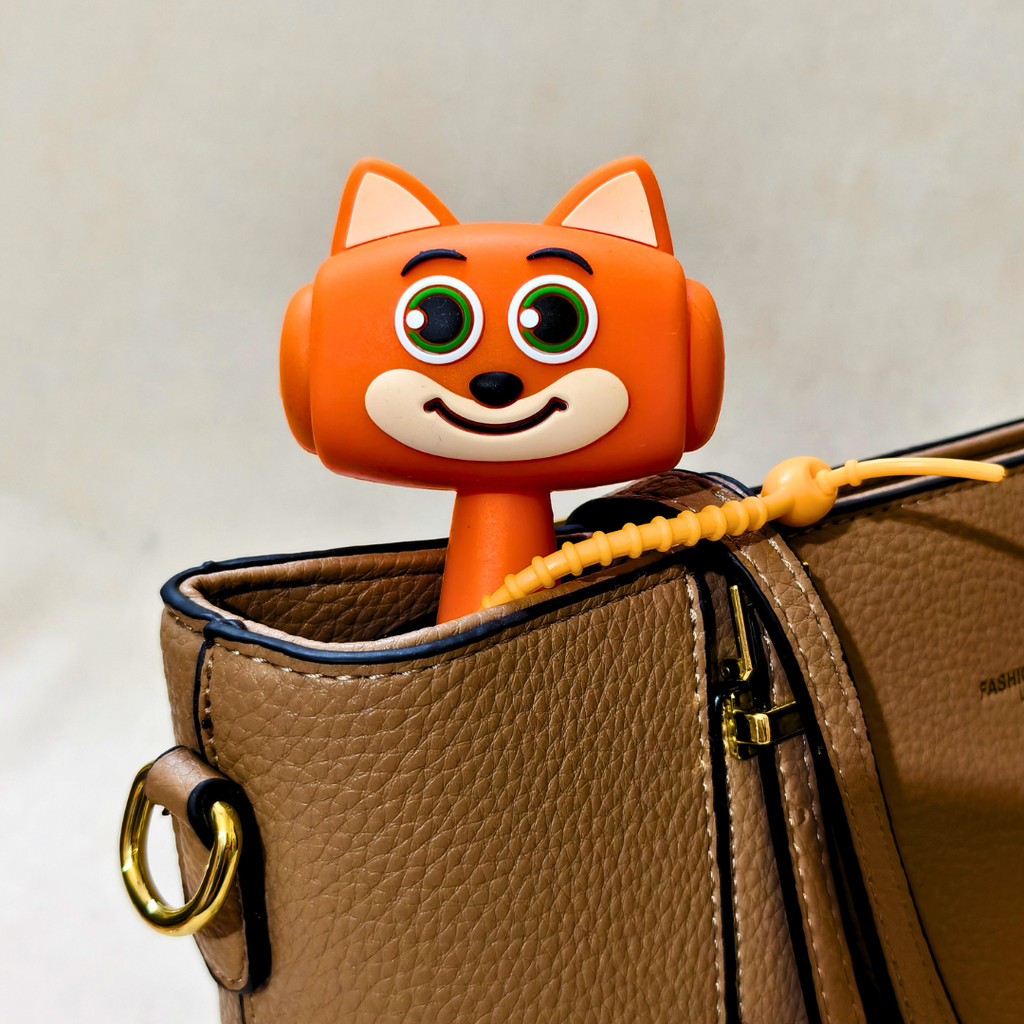 An orange cartoon fox-shaped bag charm with green eyes and a smiling mouth, inserted into the top of a brown textured leather shoulder bag with a yellow drawstring.