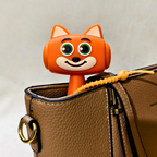 An orange cartoon fox-shaped bag charm with green eyes and a smiling mouth, inserted into the top of a brown textured leather shoulder bag with a yellow drawstring.