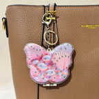 A butterfly-shaped keychain wallet with a pink and lavender marbled pattern and rhinestone embellishments, hanging from a gold-tone clasp on a brown textured handbag.