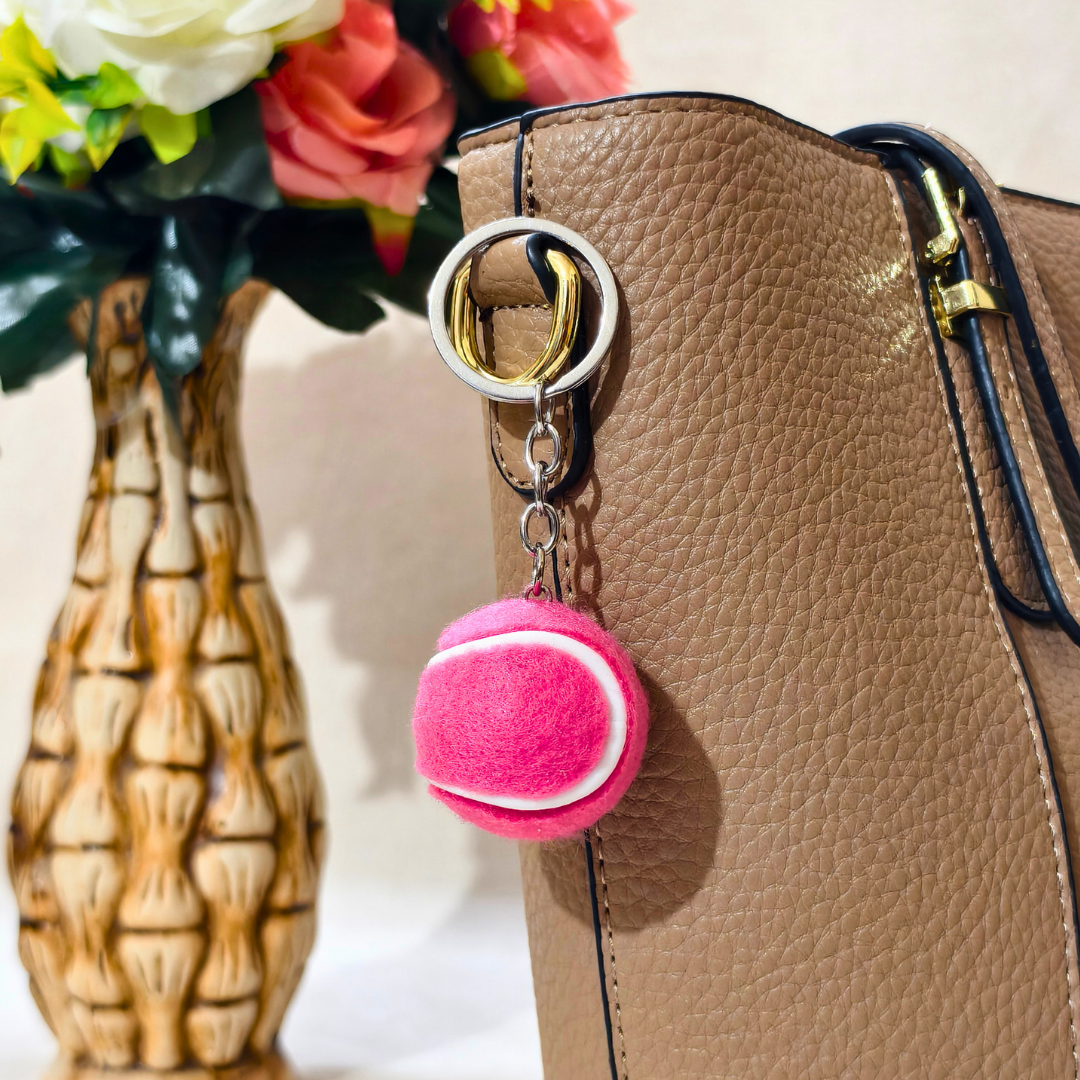 A pink tennis ball charm keychain with a silver split ring and gold keyring, hanging from a brown textured handbag next to a decorative vase with flowers.