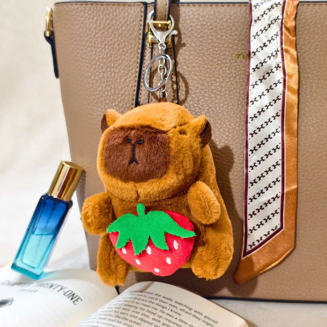 Plush bear keychain with strawberry on a handbag next to a perfume bottle and book.