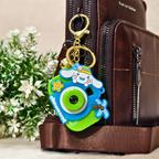 A colorful Cinnamoroll camera keychain charm with gold hardware, shown attached to a brown leather bag next to white flowers.