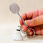 A hand holding a novelty metal keychain shaped like a badminton racket with a small white shuttleball charm, against a neutral beige background.