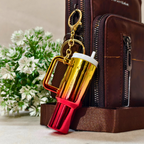 A metallic gold and red travel tumbler keychain with a white lid and gold keyring, shown leaning against a brown leather bag next to a green floral arrangement.