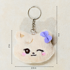 A plush keychain shaped like a cute cream-colored cat face with embroidered details, a purple bow, and a silver metal key ring, shown on a textured beige background with dimension lines indicating measurements.