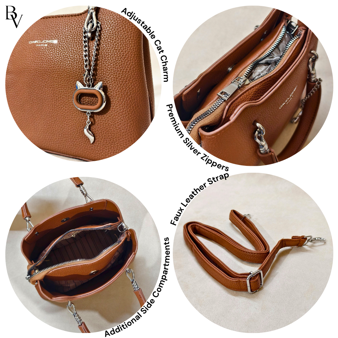 Brown leather handbag with cat charm, silver zippers, and faux leather strap on a white background.