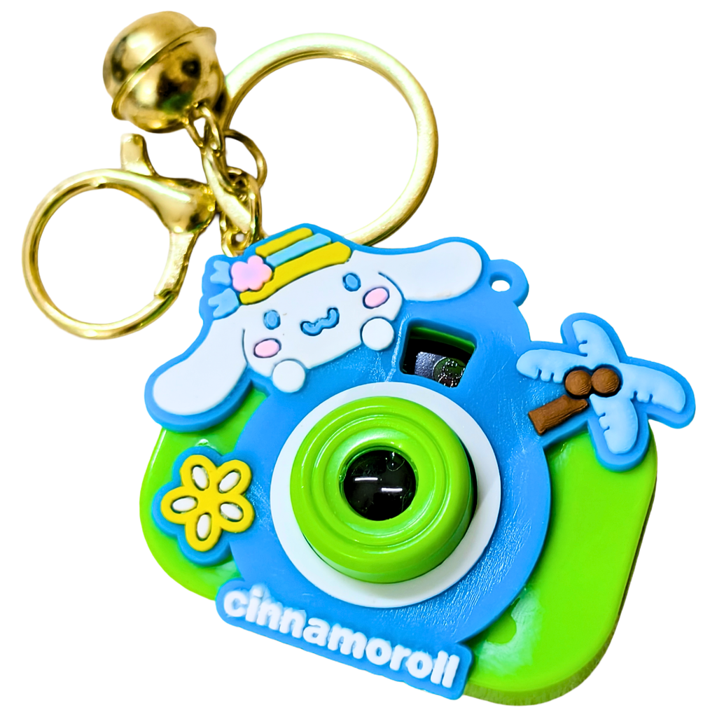 A Cinnamoroll-themed camera-shaped keychain with a gold clasp and split ring. The keychain features a blue and lime green camera body with a white Cinnamoroll character on top, a yellow flower, a palm tree, and the text 'cinnamoroll' on the front.